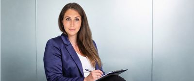 Lisa Dullnig im dunkelblauen Blazer mit Klemmbrett und Stift vor einer hellen Glaswand.